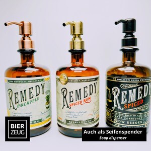 May include: Three amber glass bottles repurposed as soap dispensers with pump tops in copper, brass, and dark bronze finishes. Each bottle features a vintage-style label with the text Remedy Pineapple, Remedy Spiced Rum, and Remedy Spiced.
