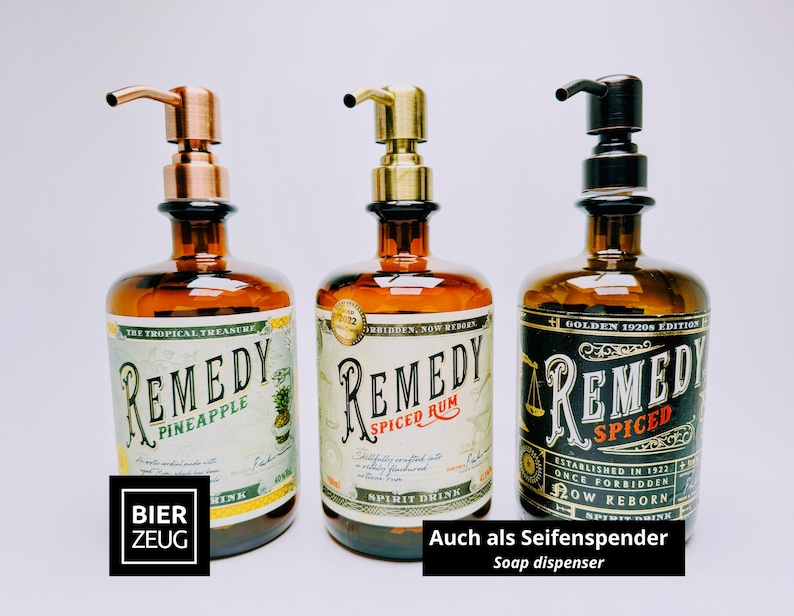 May include: Three amber glass bottles repurposed as soap dispensers with pump tops in copper, brass, and dark bronze finishes. Each bottle features a vintage-style label with the text Remedy Pineapple, Remedy Spiced Rum, and Remedy Spiced.