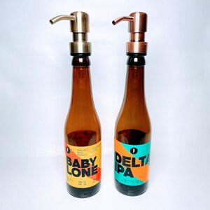 May include: Two brown glass bottles with gold and copper pump tops. The bottles have labels with the text "Baby Lone" and "Delta IPA".