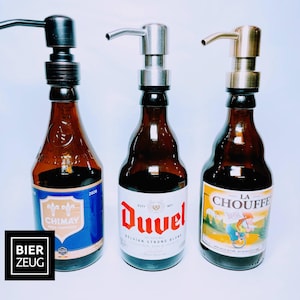 May include: Three glass bottles containing brown liquid with different coloured labels. The bottles have pump tops in black, silver and gold. The labels read "Chimay", "Duvel Belgian Strong Blond", and "La Chouffe".