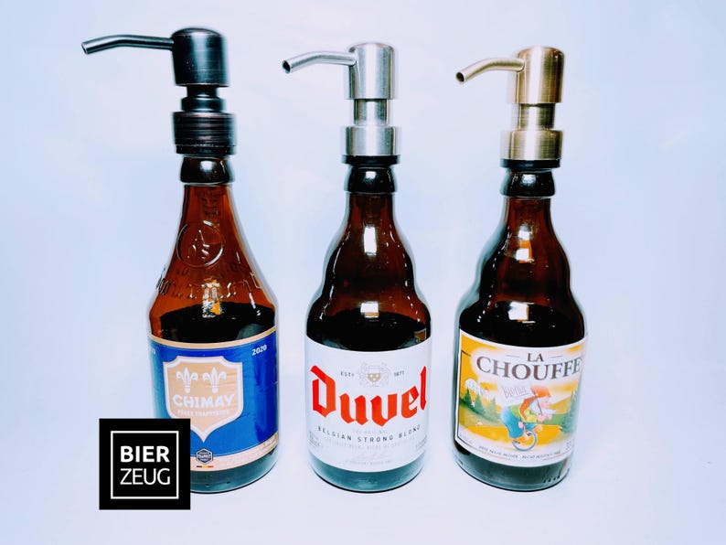 May include: Three glass bottles containing brown liquid with different coloured labels. The bottles have pump tops in black, silver and gold. The labels read "Chimay", "Duvel Belgian Strong Blond", and "La Chouffe".