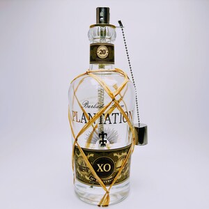 May include: A clear glass bottle of Plantation 20th Anniversary Barbados Rum wrapped in tan raffia netting. The label features the text Plantation, XO, and 20th Anniversary. A silver metal cap is attached to the neck by a small silver ball chain.