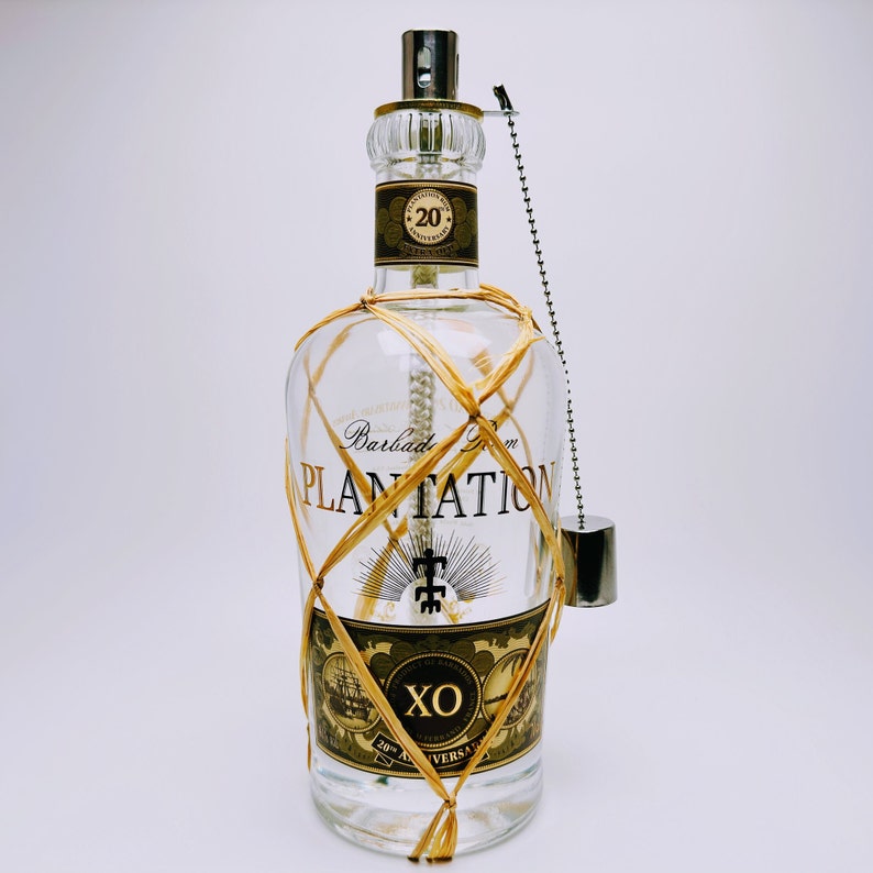 May include: A clear glass bottle of Plantation 20th Anniversary Barbados Rum wrapped in tan raffia netting. The label features the text Plantation, XO, and 20th Anniversary. A silver metal cap is attached to the neck by a small silver ball chain.