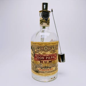 May include: A clear glass bottle of Don Papa Rum with a beige label featuring an illustration of a man, ornate gold and red text, and a metallic stopper attached by a silver ball chain. The label includes the text Don Papa Rum Small Batch.