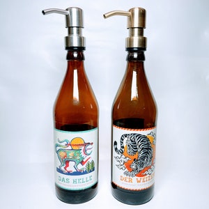May include: Two brown glass bottles with pump tops and labels. The bottle on the left has a colourful illustration of a mythical creature and the text "DAS HELLE". The bottle on the right has a black and white illustration of a tiger and the text "DER WEIZEN".