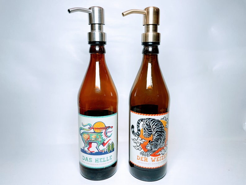 May include: Two brown glass bottles with pump tops and labels. The bottle on the left has a colourful illustration of a mythical creature and the text "DAS HELLE". The bottle on the right has a black and white illustration of a tiger and the text "DER WEIZEN".