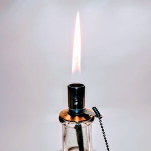 May include: A clear glass oil lamp with a burning flame extending from the top. The lamp features a metallic burner assembly with a brass-colored collar and a small black ball chain attached to a metal tab, providing a functional lighting source.