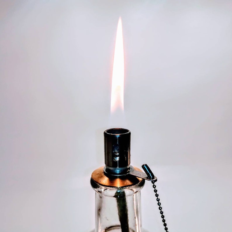 May include: A clear glass oil lamp with a burning flame extending from the top. The lamp features a metallic burner assembly with a brass-colored collar and a small black ball chain attached to a metal tab, providing a functional lighting source.