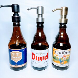 May include: Three glass bottles with different beer labels, each with a different coloured pump dispenser on top. The bottles are brown glass and the labels are colourful. The pumps are bronze, silver, and gold.