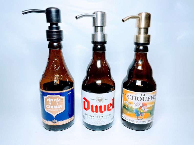 May include: Three glass bottles with different beer labels, each with a different coloured pump dispenser on top. The bottles are brown glass and the labels are colourful. The pumps are bronze, silver, and gold.