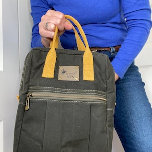 May include: A dark green canvas bag with mustard yellow handles and accents. The bag has a zippered front pocket and a small leather tag with the text "wood BEES". The bag is held by a person.