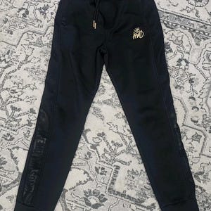May include: Black joggers featuring an elastic waistband with a drawstring, ribbed cuffs, and a gold embroidered logo on the left thigh. The sides of the legs have a vertical panel with a subtle tonal pattern and text running down the length.