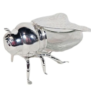 Könnte beinhalten: Ein silberfarbener, insektenförmiger dekorativer Behälter mit einem transparenten Glaskörper. Der Behälter hat detaillierte Beine, Antennen und Flügel. Die Flügel sind angelenkt und haben ein gemustertes Design. Der Behälter ist zur Aufbewahrung von Gegenständen bestimmt.
