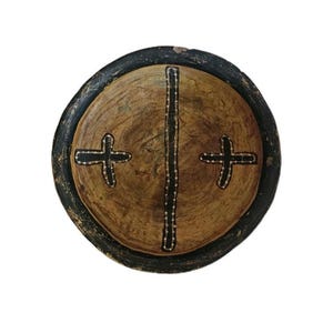 Può includere: Uno scudo rotondo in legno con un bordo marrone scuro e un centro marrone chiaro. Il centro presenta una linea nera verticale e due forme a croce nere, con una linea tratteggiata al centro.
