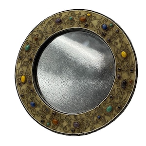 May include: A circular mirror featuring a wide, cream-coloured frame decorated with intricate gold-toned wire filigree patterns and embedded with various polished, multi-coloured oval and square cabochon stones in shades of blue, yellow, and orange.