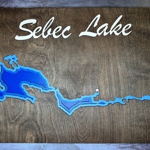 May include: A wooden sign with the words "Sebec Lake" in white lettering. The sign features a blue and white cutout of the lake.