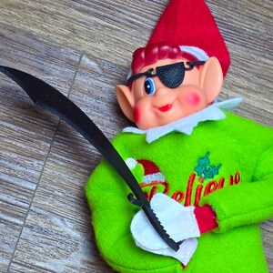 May include: A festive elf doll wearing a red and white hat, green sweater with the word "Believe", and an eye patch. The elf is holding a black sword and has red and white striped legs.