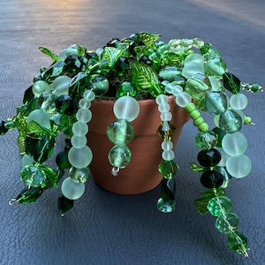 Peut inclure: Une plante décorative en perles dans un petit pot en terre cuite. La plante présente différentes nuances de perles vertes, notamment des perles rondes, facettées et en forme de feuilles, créant un effet de cascade. Le pot est posé sur une surface sombre.