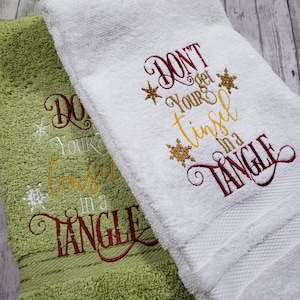 May include: Two embroidered Christmas hand towels. One is white, the other is green. Both towels feature the phrase "Don't get your tinsel in a tangle" embroidered in red, gold, and white thread, with snowflake accents. The towels are made of a soft, absorbent material.