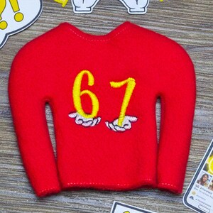 May include: A red miniature sweater with the number 67 in yellow, and the text "67: THE NEW VIBE". The image also includes cartoon hand gestures and an exclamation point.