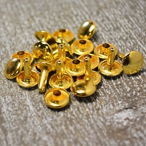 May include: A pile of gold-colored metal rivets, also known as studs or eyelets, on a wooden surface. These rivets are commonly used for leather crafting and other DIY projects.