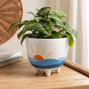 May include: A white ceramic planter with three short legs, featuring a painted design of an orange sun rising over blue ocean waves. A lush green and silver variegated pothos plant with heart-shaped leaves grows out of the top of the pot.