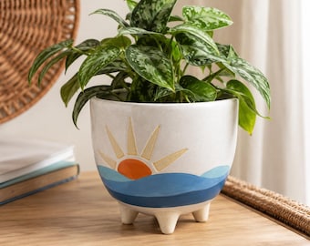 May include: A white ceramic planter with three short legs, featuring a painted design of an orange sun rising over blue ocean waves. A lush green and silver variegated pothos plant with heart-shaped leaves grows out of the top of the pot.