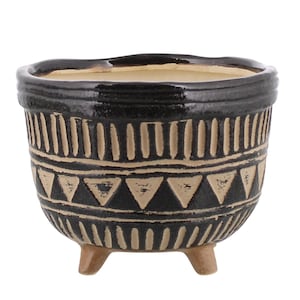 May include: A small, round ceramic planter with a glossy black glaze and a cream-colored interior. The exterior features a hand-carved pattern of vertical lines and triangles in a light beige tone. The planter stands on three short, unglazed brown feet.