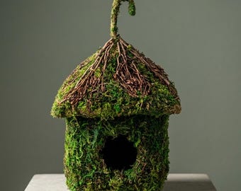 May include: A decorative birdhouse covered in green moss, with a brown twig roof and a curved handle. The birdhouse has a circular opening and sits on a gray surface. The overall design is natural and rustic.
