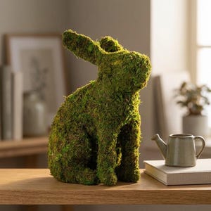May include: A decorative moss-covered rabbit sculpture in a seated position. The green rabbit has long ears and is placed on a wooden surface. A small, gray watering can and a book are next to the rabbit.