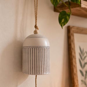 May include: A ceramic bell with a white finish and black vertical line detailing. The bell features a rounded top with a single wooden bead and hangs from a tan jute rope. A matching jute rope extends from the center of the bell as a clapper.