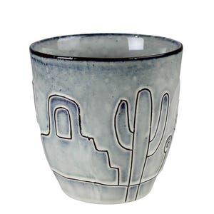 May include: A ceramic mug with a light blue and white mottled glaze finish. The exterior features a dark, hand-drawn style outline of a desert landscape, including a cactus and a building structure, etched into the surface of the vessel.