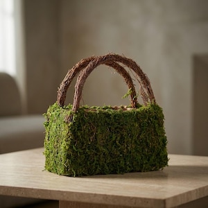 May include: A rectangular basket covered in vibrant green moss, featuring two arched handles made of intertwined brown twigs. The basket is set on a light-colored surface, suggesting a natural, decorative theme.