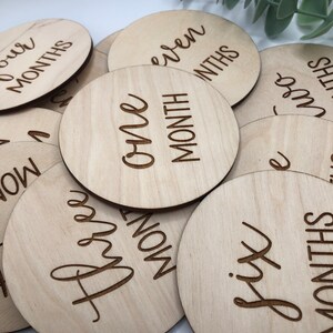 May include: A collection of round, light-colored wooden discs with engraved text indicating baby's age in months. The discs are arranged in a slightly overlapping pattern, with the text "One Month" clearly visible.