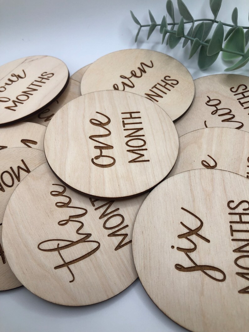 May include: A collection of round, light-colored wooden discs with engraved text indicating baby's age in months. The discs are arranged in a slightly overlapping pattern, with the text "One Month" clearly visible.