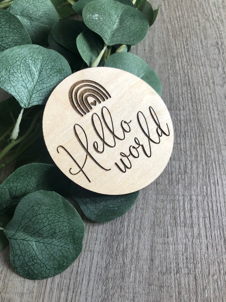 May include: A round wooden disc with the words "Hello world" in a cursive font. Above the text is a small engraved rainbow design. The disc is light-colored and sits on a background of green leaves and a gray wooden surface.