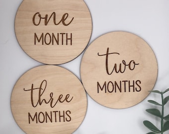 May include: Three round wooden discs with engraved text. The discs are light brown and feature the words "one MONTH", "two MONTHS", and "three MONTHS" in a script font. The discs are likely baby milestone markers.