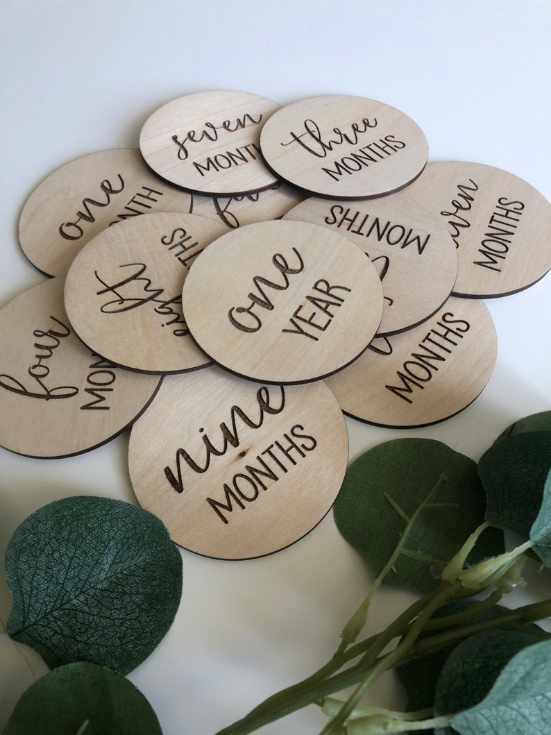 May include: A collection of wooden baby milestone discs. Each disc is round and engraved with a month or year, such as "one month," "three months," and "one year." The discs are arranged on a white surface with green foliage.