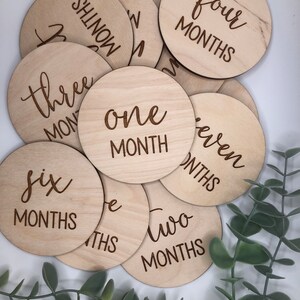May include: A collection of wooden, circular milestone markers. Each disc is engraved with a month number, from one to seven months, in a stylish script font. The natural wood grain provides a warm, neutral tone, perfect for baby photos.