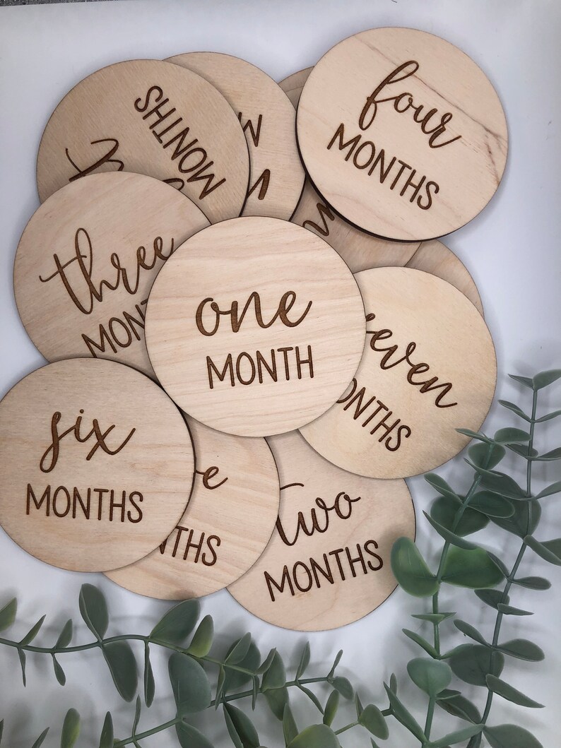 May include: A collection of wooden, circular milestone markers. Each disc is engraved with a month number, from one to seven months, in a stylish script font. The natural wood grain provides a warm, neutral tone, perfect for baby photos.
