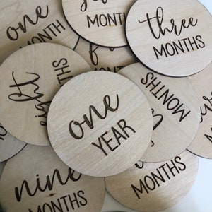 May include: A collection of light wood, circular milestone markers with laser-etched black text. The markers display phrases like "one month," "three months," and "one year." The wood grain is visible, and the overall aesthetic is minimalist and natural.
