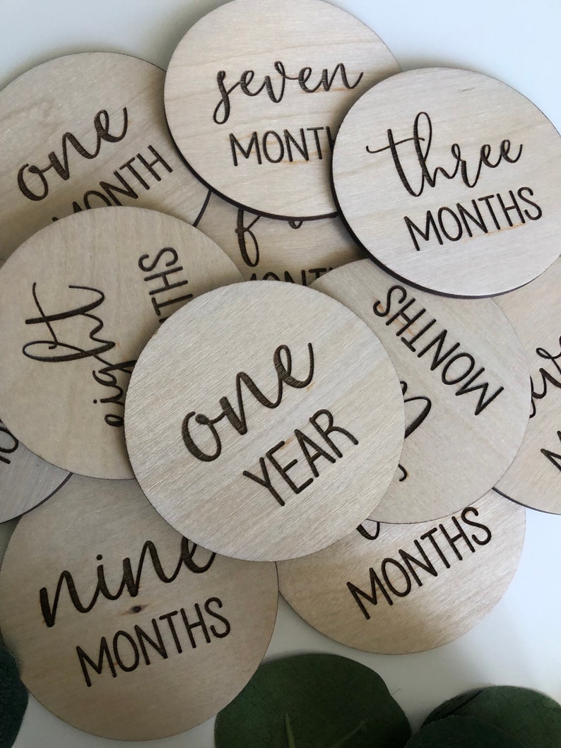 May include: A collection of light wood, circular milestone markers with laser-etched black text. The markers display phrases like "one month," "three months," and "one year." The wood grain is visible, and the overall aesthetic is minimalist and natural.