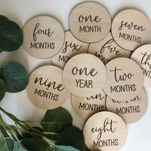 May include: A collection of wooden milestone discs with engraved text, including "one month," "two months," and "one year." The discs are arranged on a white surface, with green foliage in the background. These are baby milestone markers.