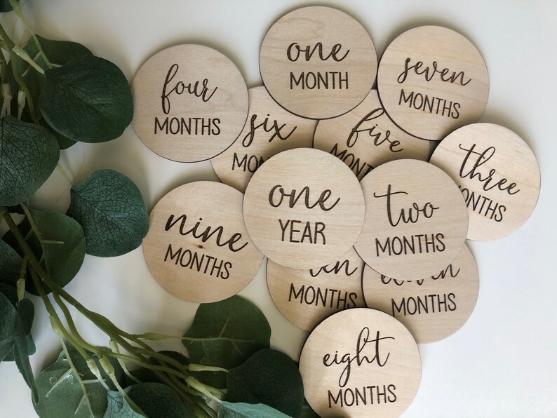 May include: A collection of wooden milestone discs with engraved text, including "one month," "two months," and "one year." The discs are arranged on a white surface, with green foliage in the background. These are baby milestone markers.