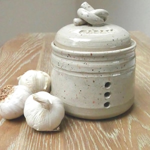 May include: A cream-coloured ceramic garlic keeper with a knotted handle on the lid. The word "GARLIC" is etched on the lid. Three garlic bulbs are next to the keeper on a wooden surface.