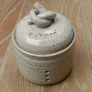 May include: A speckled ceramic garlic keeper with a knotted handle on the lid. The word "GARLIC" is embossed on the lid. The container has three small holes on the side and is beige with black and red speckles.