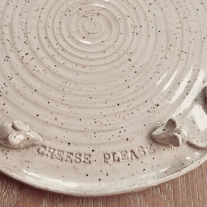 May include: A speckled, cream-coloured ceramic plate with a spiral design and three raised mouse figures. The words "CHEESE PLEASE" are embossed on the plate's surface. A whimsical, decorative serving dish.