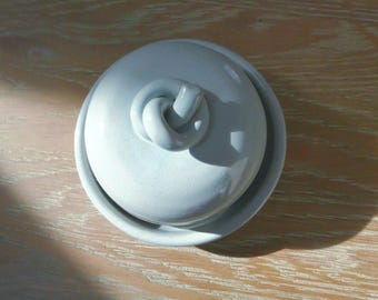 May include: A round, light gray ceramic butter dish with a matching lid. The lid features a sculptural, knotted handle at the center. The smooth, glossy finish reflects light, highlighting the circular shape of the base and the domed cover.