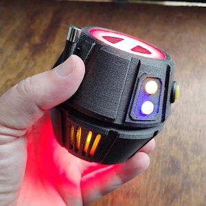 May include: A black, textured, cylindrical electronic prop with a red glowing top, a small silver antenna, and side panels featuring one yellow and one blue light. The base of the device emits a bright red light through vertical vents.