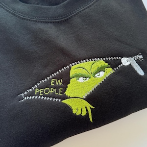 May include: A black crewneck sweatshirt featuring an embroidered design of a green character peeking through a zippered opening. The embroidery includes the text "EW. PEOPLE" in yellow lettering, with the character looking down with a frown.